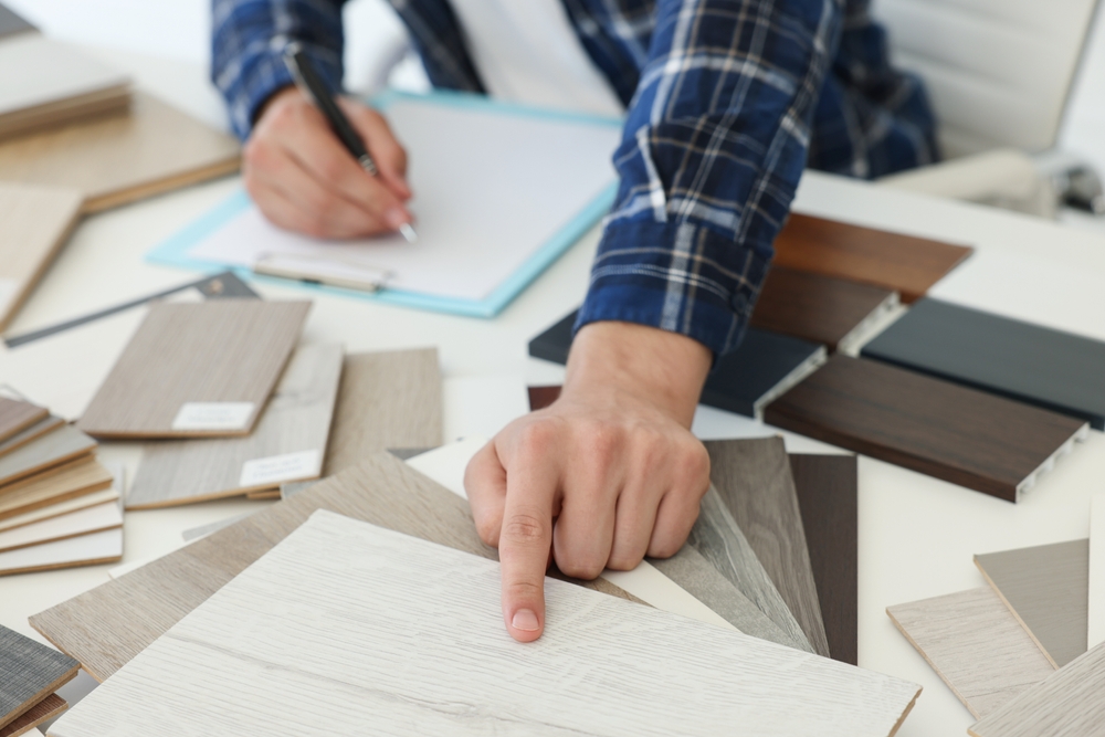 A close-up shot of a contractor reviewing flooring, cabinet, and hardware samples on a table. Include hands, tape measure, or plans to show “real project planning.